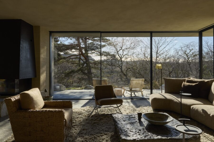 A sitting room at Hope, with a curved sofa, woven chairs and glass walls opening onto a terrace and trees